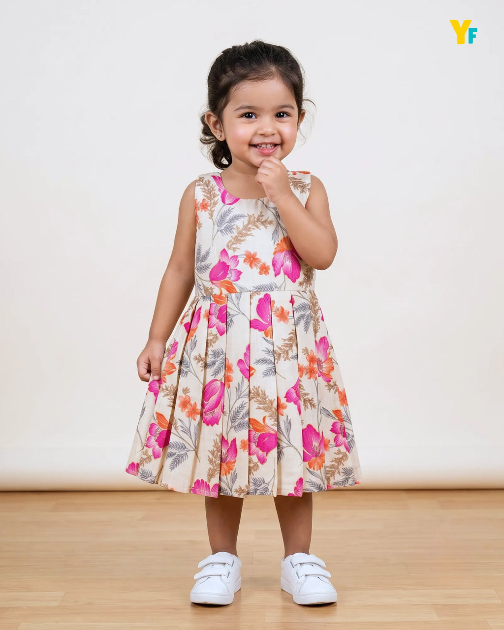 A young girl stands smiling in a sleeveless floral dress with a pleated skirt, paired with white sneakers, against a plain cream wall and light wood floor.