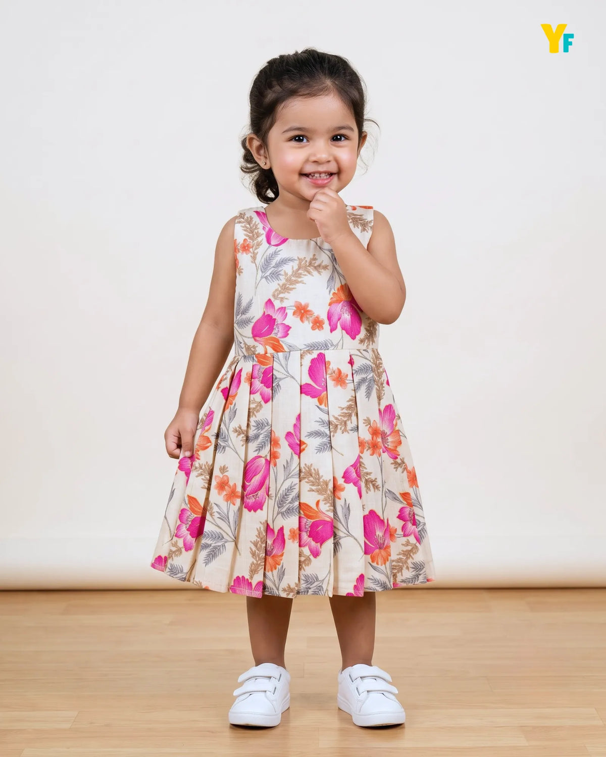 A young girl stands smiling in a sleeveless floral dress with a pleated skirt, paired with white sneakers, against a plain cream wall and light wood floor.